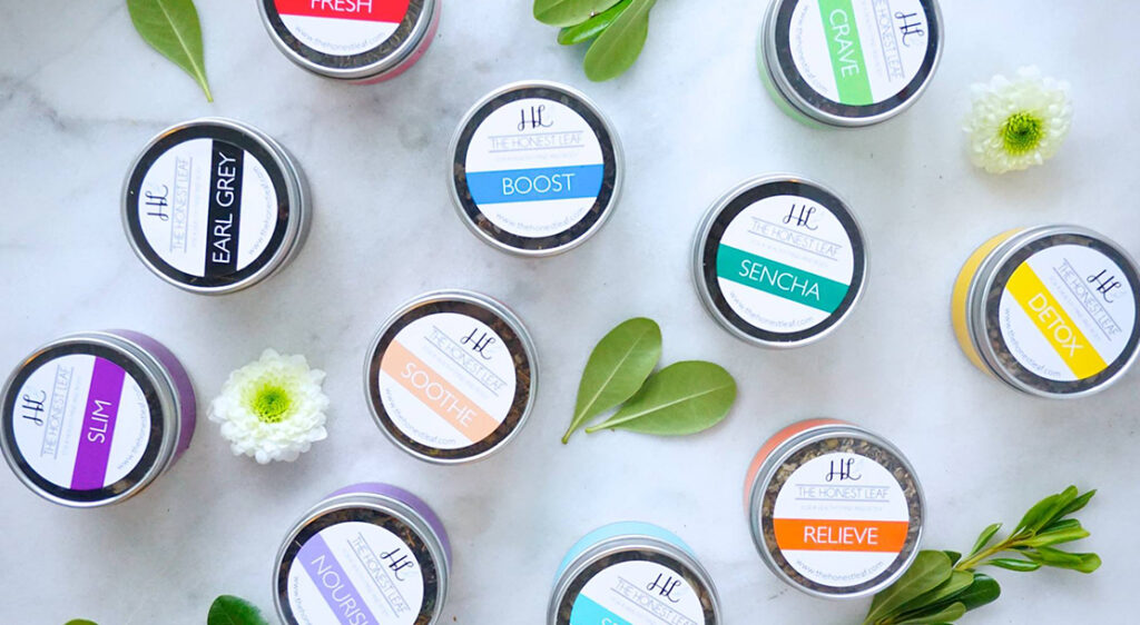 Photograph of round tea tins scattered on a white-marble counter top with colourful labels of the tea names of Honest Leaf blends as follows Earl Grey, Slim, Sencha, Boost, Sooth, Nourish, Relieve, Detox, and Crave. 